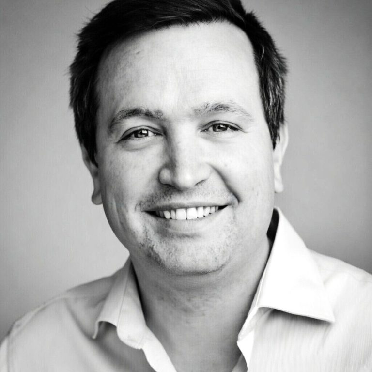 Retrato de un hombre sonriendo, con una camisa clara y fondo blanco.